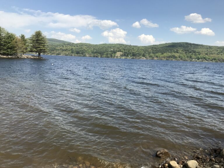 Great Sacandaga Lake with rippled water, a rocky foreground, and wooded hills along the opposite shore.