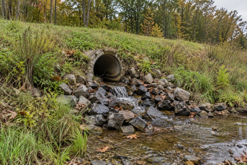 Illustrative concrete drainage culvert with water flowing over stones beside an autumn woodland.